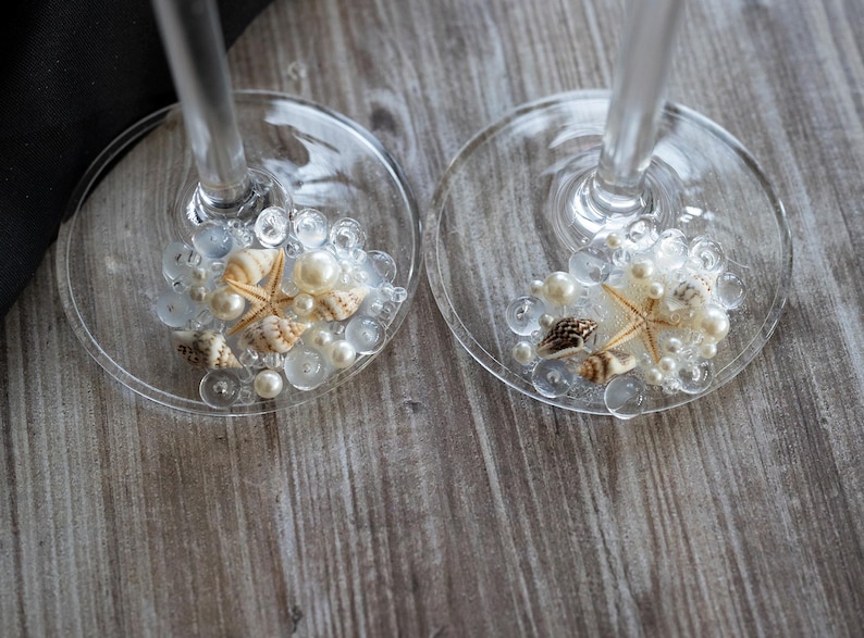 May include: Two clear glass wine glasses with white pearl beads and seashells inside. The glasses are on a wooden surface.
