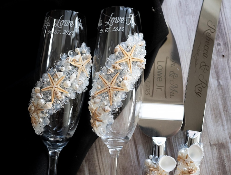 May include: Two champagne flutes decorated with seashells and starfish, with the text "Mr. Lowe Jr. 06.07.2025" and "Mrs. Lowe Jr. 06.07.2025" engraved on the glasses. A silver cake knife with the text "Rebecca & Jay Mr. & Mrs. Lowe Jr. 06.07.2025" is also included.