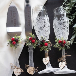 May include: A wedding cake cutting set with two champagne flutes. The set is decorated with sprigs of greenery and red berries. The cake knife and flutes are engraved with the couple's names and wedding date. The flutes have a white lace design.