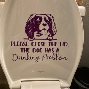 Peut inclure: Abattant de toilette blanc avec un dessin violet d'un chien et le texte "PLEASE CLOSE THE LID, THE DOG HAS A Drinking Problem". Le dessin du chien est un Cavalier King Charles Spaniel.