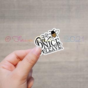 May include: A white sticker with the words "Bee Nice or Leave" in a playful font, accompanied by a black and yellow bee illustration. The sticker is held in a hand against a blurred wooden backdrop.