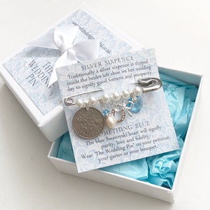Puede incluir: Una caja blanca con un forro estampado en azul y blanco contiene un alfiler de boda de seis peniques de plata. El alfiler está adornado con perlas, un corazón de cristal Swarovski azul, un amuleto de herradura y un pequeño amuleto azul. La caja incluye una tarjeta con el texto "The Wedding Pin" e instrucciones sobre cómo usar el alfiler.