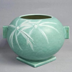 May include: A round, light green ceramic vase with a matte finish, featuring two side handles and a square base. The exterior is decorated with a raised, white, stylized floral pattern of thin, pointed petals radiating from a central point.