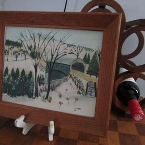 May include: A framed painting of a winter landscape featuring a covered bridge, snow-covered ground, and evergreen trees. The artwork is enclosed in a medium-toned wooden frame and is signed with the initials IMS in the bottom right corner.