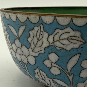 Może przedstawiać: Zbliżenie dekoracyjnej miski z jasnoniebieskim tłem i białym kwiatowym wzorem. Obręcz jest karbowana ze złotym wykończeniem. Wnętrze jest zielone.