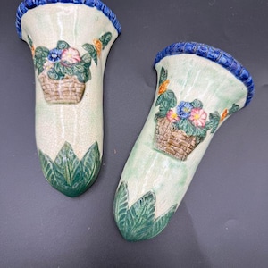 May include: Two ceramic wall pockets featuring a cream-coloured crackle glaze finish. Each piece has a blue scalloped rim, a raised relief design of a woven basket filled with colourful flowers, and green leaves at the pointed base of the vessel.
