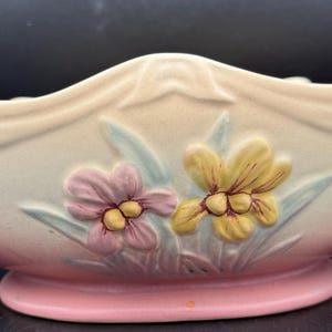 May include: A ceramic planter in a boat shape, with a pink base and a cream-colored body. The planter features embossed flowers in pink and yellow, with green leaves. The rim has a decorative, draped design.
