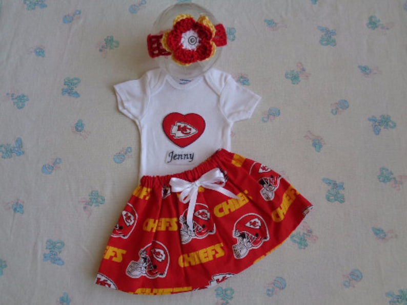 May include: A baby outfit set featuring a white onesie with a red heart-shaped patch and the name "Jenny" embroidered below. The set includes a red skirt with a white bow and a matching headband, all adorned with a Chiefs football team design.