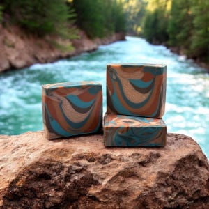 May include: Three bars of handmade soap with a swirling pattern of brown, blue, and white. The soap is sitting on a rock with a river in the background.