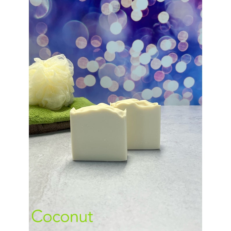 K&ouml;nnte beinhalten: Zwei wei&szlig;e, quadratische Seifenst&uuml;cke mit strukturierter Oberseite. Ein gelber Luffa und ein gr&uuml;nes Handtuch befinden sich im Hintergrund. Das Wort "Coconut" steht gr&uuml;n am unteren Bildrand.