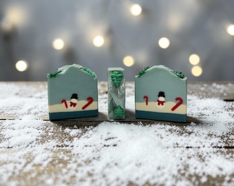 May include: Three rectangular bars of soap arranged upright. The two outer bars feature a light blue base with a white snowy ground, a small snowman with a black hat, and red candy canes. The center bar is grey with green tree accents.