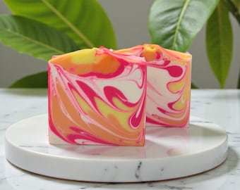 Op de afbeelding: Twee vierkante stukken handgemaakte zeep met een levendig wervelontwerp. De zeep heeft een mix van kleuren, waaronder roze, geel, oranje en wit. De zepen worden gepresenteerd op een witte marmeren plaat, met groene bladeren op de achtergrond.