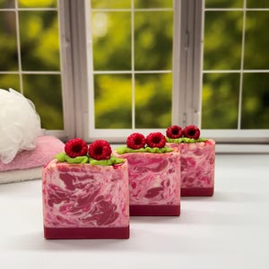 May include: Three square soaps with a raspberry design. The soaps have a pink and white marbled pattern with a dark pink base. Each soap is topped with a green leaf design and three red raspberries. The soaps are on a white surface.