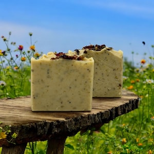 May include: Two bars of handmade soap with dried flower petals on top. The soap is a light beige color and has a grainy texture. The soap is sitting on a piece of wood with a blurred background of wildflowers.