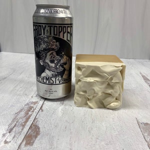 May include: A silver can of "Heady Topper" ale from The Alchemist brewery in Vermont. The can features a cartoon illustration of a man with a beard drinking from a glass. The text on the can reads "Heady Topper" and "The Alchemist - Vermont". The can is sitting on a white wooden surface next to a stack of three white and beige foam blocks.