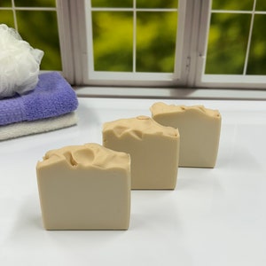 K&ouml;nnte beinhalten: Drei beige quadratische Seifen sind in einer Reihe angeordnet. Die Seifen haben eine leicht strukturierte Oberfl&auml;che. Ein Stapel gefalteter Handt&uuml;cher und ein wei&szlig;er Badeschwamm befinden sich im Hintergrund. Die Szene spielt sich vor einem Fenster mit gr&uuml;ner Au&szlig;enansicht ab.