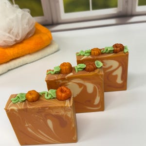 May include: Three rectangular bars of soap with a marbled orange and cream swirl pattern. Each bar is topped with small, sculpted orange pumpkins and light green leaves made of soap, arranged along the top edge of the flat surface.