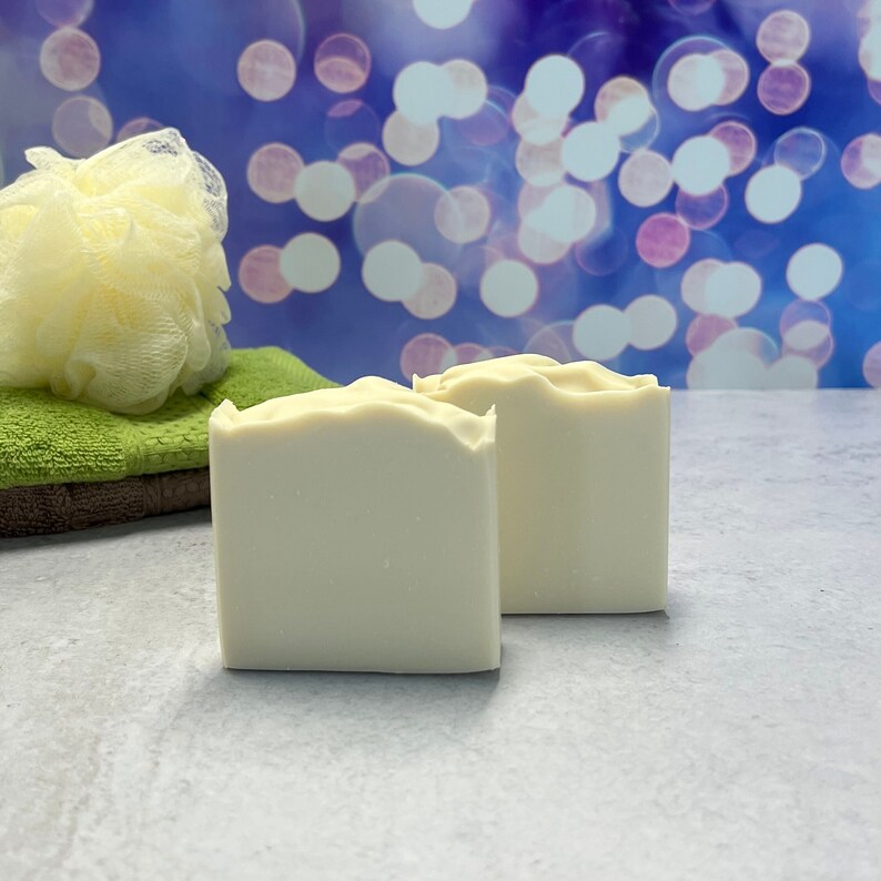 K&ouml;nnte beinhalten: Zwei quadratische, cremefarbene Seifenst&uuml;cke mit gewelltem oberem Rand. Ein gr&uuml;nes Waschtuch und ein gelber Luffa befinden sich im Hintergrund. Der Hintergrund ist ein verschwommenes Blau mit wei&szlig;en Bokeh-Lichtern.