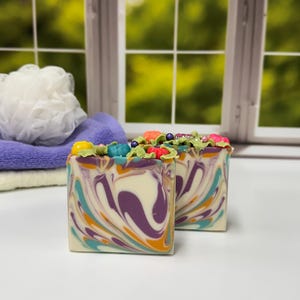 May include: Two rectangular bars of handcrafted soap featuring a marbled swirl pattern in shades of purple, teal, orange, and white. The tops are decorated with small, colorful sculpted shapes resembling berries and leaves in bright hues.
