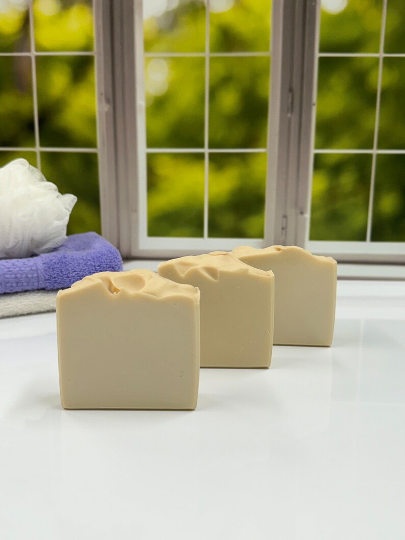 K&ouml;nnte beinhalten: Drei beige, quadratische Seifenst&uuml;cke sind in einer Reihe angeordnet. Die Seifen haben eine leicht strukturierte Oberfl&auml;che. Im Hintergrund ist ein Fenster mit verschwommener gr&uuml;ner Au&szlig;enansicht zu sehen, sowie ein Stapel gefalteter Handt&uuml;cher und ein wei&szlig;er Badeschwamm.