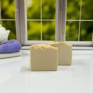 K&ouml;nnte beinhalten: Zwei beige quadratische Seifenst&uuml;cke werden auf einer wei&szlig;en Oberfl&auml;che pr&auml;sentiert, mit einem Fenster und Gr&uuml;n im Hintergrund. Links sind ein Stapel gefalteter Handt&uuml;cher und ein Badeschwamm zu sehen. Die Seifen haben eine strukturierte Oberseite.