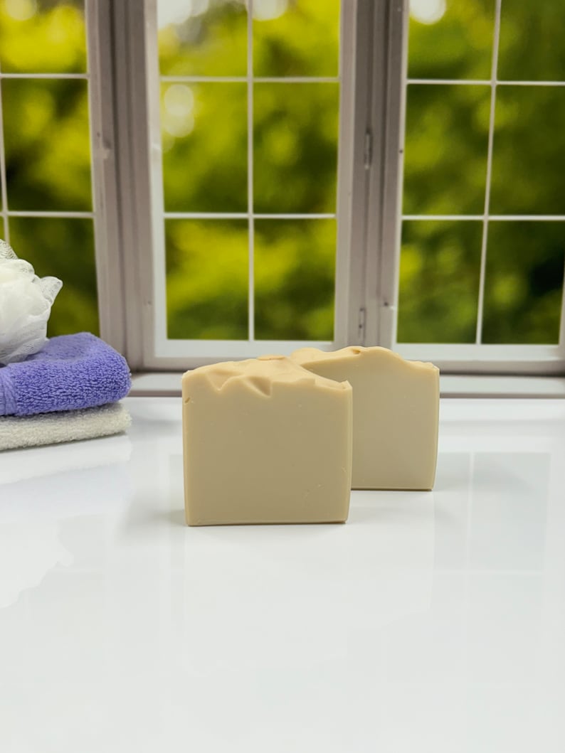 K&ouml;nnte beinhalten: Zwei beige quadratische Seifenst&uuml;cke werden auf einer wei&szlig;en Oberfl&auml;che pr&auml;sentiert, mit einem Fenster und Gr&uuml;n im Hintergrund. Links sind ein Stapel gefalteter Handt&uuml;cher und ein Badeschwamm zu sehen. Die Seifen haben eine strukturierte Oberseite.