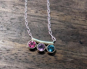 Puede incluir: Un collar de plata con tres piedras preciosas de colores: rosa, morado y turquesa. Las piedras están engarzadas en un colgante de barra curvada, suspendido de una delicada cadena. El collar se muestra sobre una superficie de madera oscura.