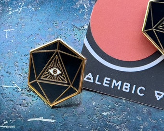 Puede incluir: Un pin de esmalte negro y dorado con un diseño geométrico y un ojo que todo lo ve en el centro. El pin tiene forma de dado de 20 caras.