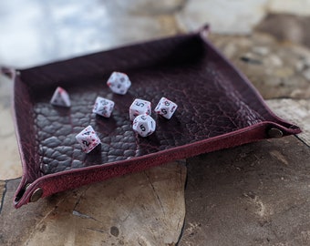 Puede incluir: Un tapete para dados de cuero burdeos con dados blancos con números negros y salpicaduras de pintura roja. El tapete para dados tiene un borde cosido y una solapa.