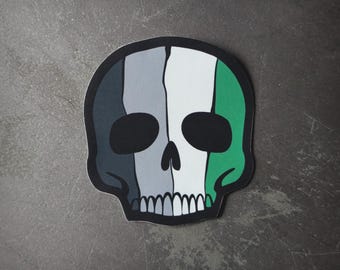 Puede incluir: Una pegatina de calavera en blanco y negro con rayas verdes, grises y blancas. La calavera tiene un diseño simple con ojos negros y una nariz blanca.