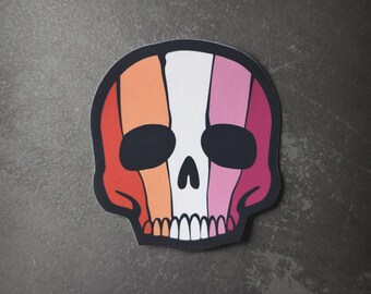 Puede incluir: Una pegatina con forma de calavera con un contorno negro. La calavera está coloreada con rayas verticales rojas, naranjas, blancas, rosas y granates. Las cuencas de los ojos son negras y los dientes blancos.