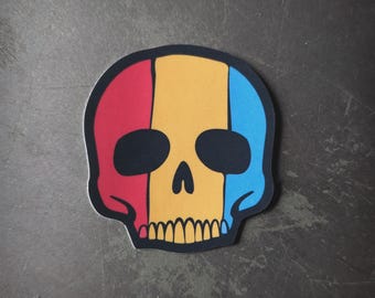 Puede incluir: Una pegatina de calavera negra con rayas rojas, amarillas y azules. La calavera está delineada en negro y tiene un diseño simple.