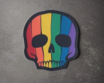 Puede incluir: Una pegatina de calavera arcoíris con contorno negro. La calavera está rayada con rojo, naranja, amarillo, verde, azul, añil y violeta.