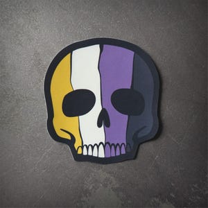 Peut inclure: Un autocollant de crâne noir et blanc avec un schéma de couleurs jaune, blanc et violet. Le crâne est stylisé avec un design simple et est un symbole populaire de la communauté non binaire.