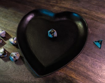 Puede incluir: Una bandeja de dados de corazón negro con un dado iridiscente azul y dorado en la parte superior. Hay varios dados más esparcidos alrededor de la bandeja sobre una superficie de madera.