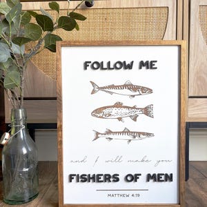 May include: Wooden framed art print with the text "FOLLOW ME" and "FISHERS OF MEN" in black 3D letters. The print features three fish illustrations in brown and the Bible verse Matthew 4:19.