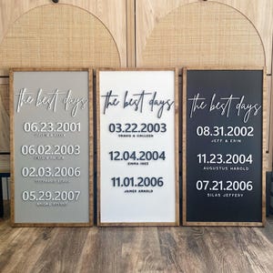 May include: Three framed wooden signs with the text "the best days" in cursive. Each sign has a different background color: gray, white, and black. Each sign displays dates and names, likely commemorating special occasions.