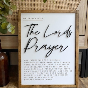 Może przedstawiać: Biały drewniany znak z brązową ramą zawiera tekst "The Lord's Prayer" w czarnym, ozdobnym piśmie. Tekst modlitwy Pańskiej jest wydrukowany czarnym kolorem pod tytułem, a na g&oacute;rze znajduje się odniesienie do Pisma Świętego "Matthew 6:10-13".