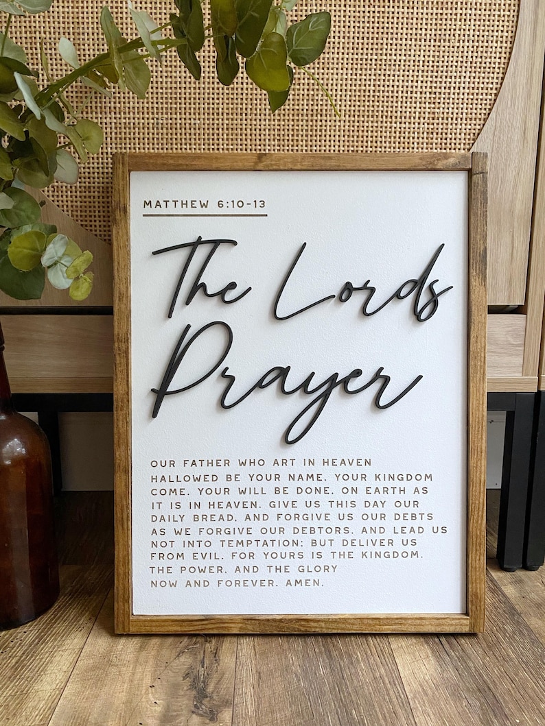 Może przedstawiać: Biały drewniany znak z brązową ramą zawiera tekst "The Lord's Prayer" w czarnym, ozdobnym piśmie. Tekst modlitwy Pańskiej jest wydrukowany czarnym kolorem pod tytułem, a na g&oacute;rze znajduje się odniesienie do Pisma Świętego "Matthew 6:10-13".
