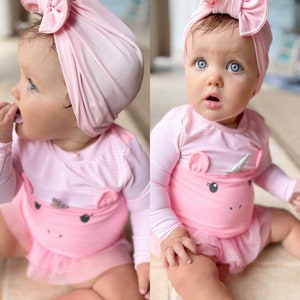 May include: A baby wearing a pink swimsuit with a unicorn design and a matching pink bow headband. The swimsuit has long sleeves and a tutu-style skirt. The baby has blue eyes.