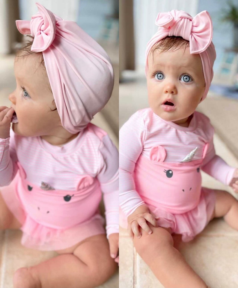 May include: A baby wearing a pink swimsuit with a unicorn design and a matching pink bow headband. The swimsuit has long sleeves and a tutu-style skirt. The baby has blue eyes.