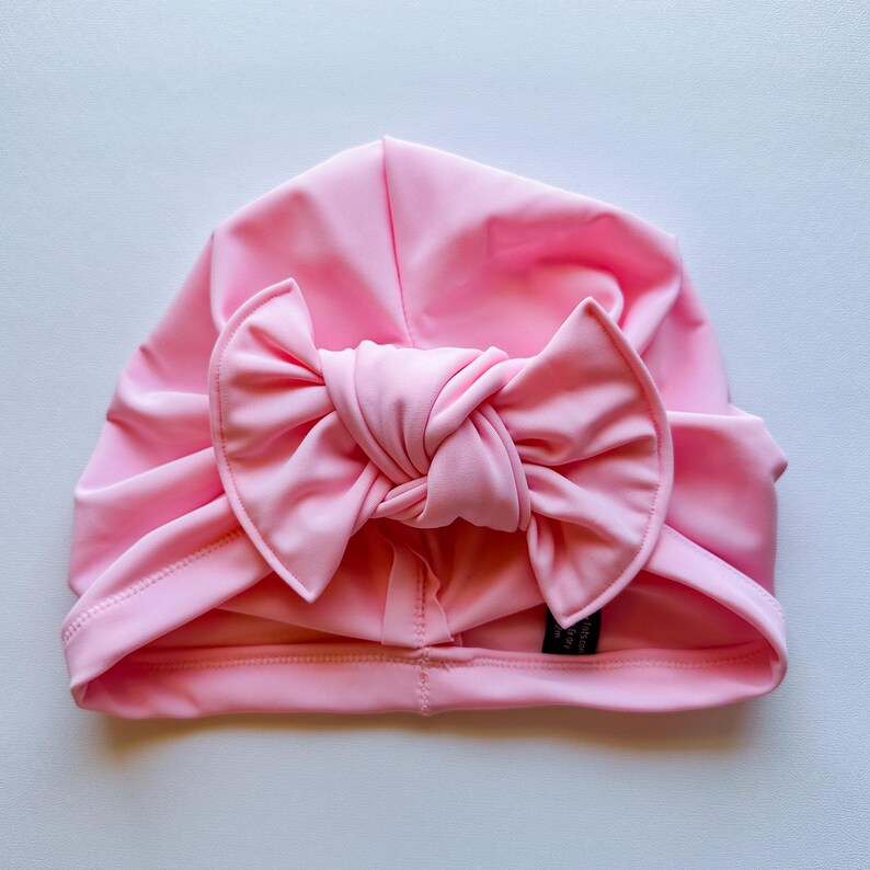 May include: A light pink turban-style baby hat with a large bow on the front. The hat is made of a soft, stretchy material and is designed to fit snugly on the head. The bow is a prominent feature, adding a touch of elegance.