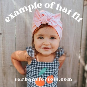 May include: A young child wearing a light pink turban with a large bow. The child is also wearing a black and white checkered dress with a cherry design. The text "example of hat fit" and "turbansfortots.com" are visible.