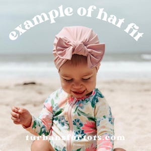 May include: A baby wearing a light pink turban with a large bow. The text "example of hat fit" is arched above the baby's head. The baby is wearing a floral print swimsuit and is sitting on a sandy beach.