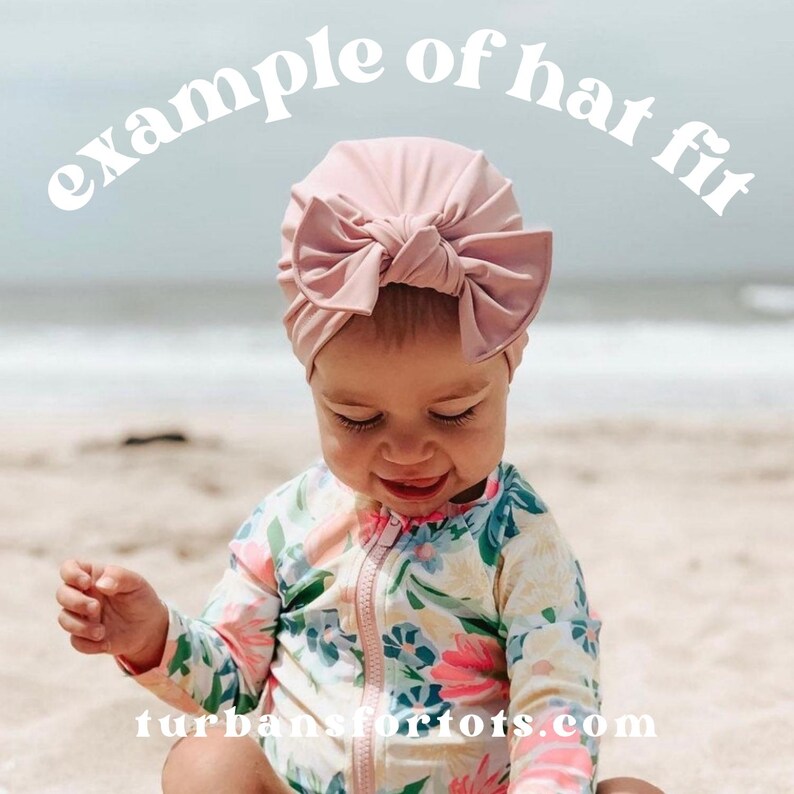 May include: A baby wearing a light pink turban with a large bow. The text "example of hat fit" is arched above the baby's head. The baby is wearing a floral print swimsuit and is sitting on a sandy beach.