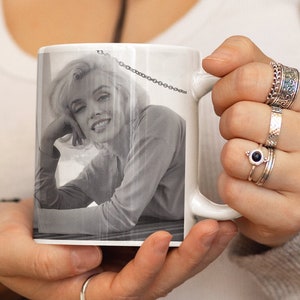 May include: A white ceramic mug featuring a black and white photograph of Marilyn Monroe. The mug is being held by a person wearing a light brown cardigan and a white vest top.