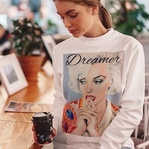 May include: White long-sleeve sweatshirt with a graphic print of Marilyn Monroe. The print features the text "Dreamer" above her image. The sweatshirt has a crew neck and long sleeves.