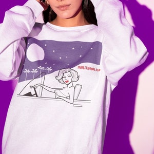 Puede incluir: Sudadera blanca con un estampado gráfico morado y blanco de una mujer conduciendo un coche por la noche con el texto "HOLLYWOODLAND" en la esquina superior derecha.
