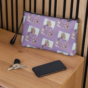 Puede incluir: Una cartera de mano de color morado con una cremallera negra y una correa negra. La cartera tiene un patrón repetido de una mujer con un vestido morado.