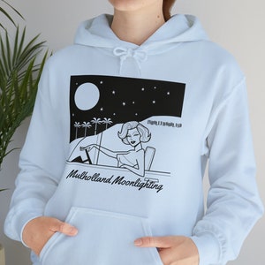Puede incluir: Una sudadera con capucha azul claro con una ilustración en blanco y negro de una mujer conduciendo un coche por la noche con palmeras y una luna llena en el fondo. El texto "Mulholland Moonlighting" está impreso debajo de la ilustración.